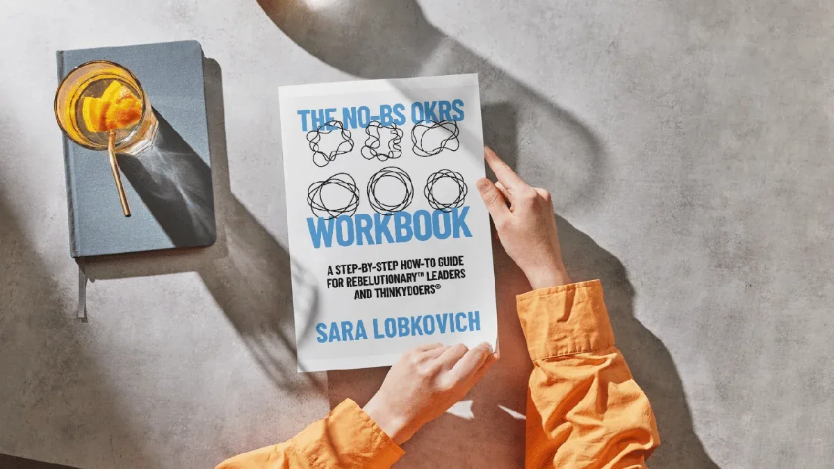 Hands holding The No-BS OKRs Workbook with coffee and journal nearby